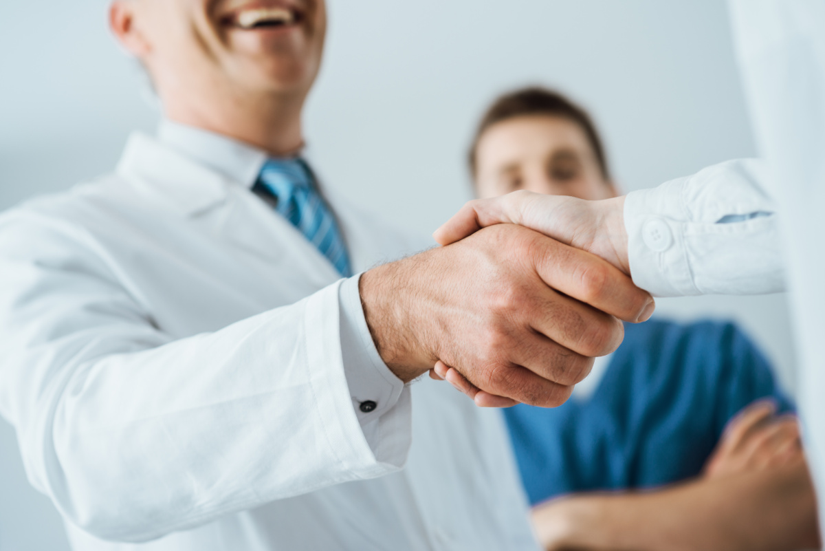 A doctor shaking hands with someone with a nurse corssing his arms in the background